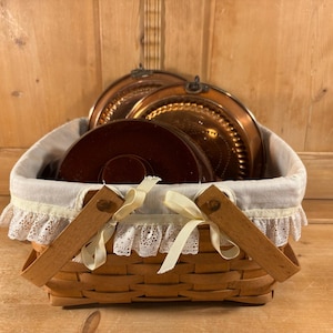 May include: A woven basket with a white fabric lining and lace trim, holding copper plates and a dark brown plate. The basket has wooden handles tied with cream-colored ribbons. The background is a wooden surface.