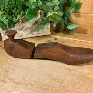 May include: A vintage brown wooden shoe last featuring a two-piece construction with a metal hinge mechanism connecting the heel and toe sections, and a small cylindrical knob located on the top of the heel for easy removal from footwear.
