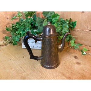 May include: A vintage brass coffee pot with a black wooden handle. The pot has a swirl design and a lid with a knob.