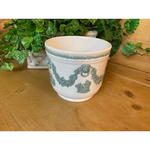May include: A white ceramic planter with a green embossed design of grapevines and a lion's head. The planter has a round shape and a wide rim.