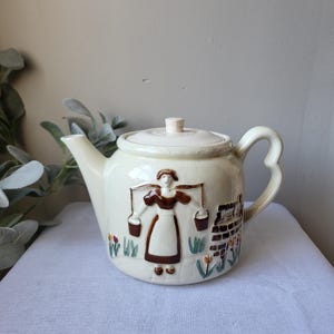 May include: A cream-colored ceramic teapot with a brown and white illustration of a woman carrying buckets. The teapot has a brown handle and a brown lid.