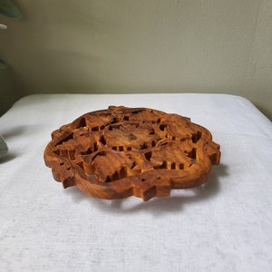 May include: A round, wooden coaster with a carved design of intricate floral patterns. The coaster is made of dark brown wood and has a smooth, polished finish.