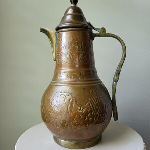 May include: A copper pitcher with a lid, featuring an etched camel and floral design on the body. The vessel has a curved handle, a pointed spout, and a rounded base, showcasing a weathered metallic finish with visible patina and dark accents.