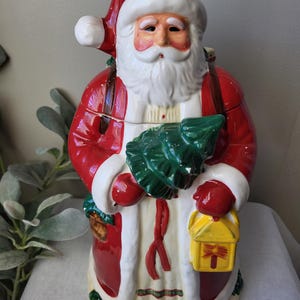 May include: A ceramic Santa Claus figure, painted in glossy red, white, and cream colors. Santa is holding a green Christmas tree and a yellow lantern. The figure has a white beard and a red hat with a white pom-pom.