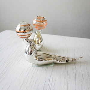 May include: Two silver glass ornaments shaped like birds, each balancing a round, orange and silver striped ball on their beaks.