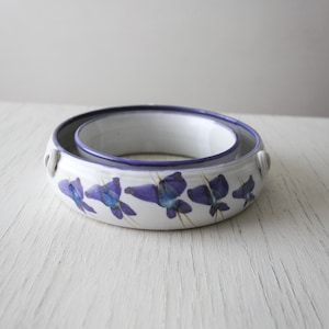 May include: Two-piece ceramic bowl set. The bowls are white with a purple floral pattern and a purple rim. The larger bowl has two small handles. The set is on a white surface.