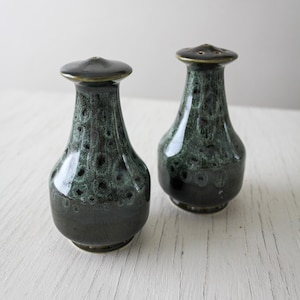 May include: A pair of vintage ceramic salt and pepper shakers. The shakers are dark green with a speckled pattern and a glossy finish. The tops are dark with small holes for dispensing. The shakers have a bulbous shape and sit on a white surface.