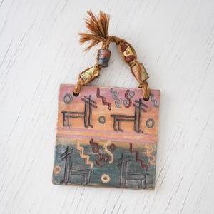 May include: A rectangular ceramic wall hanging with a painted design featuring stylized animal figures and geometric patterns in shades of pink, orange, blue, and green. It has a braided rope hanger with decorative beads.