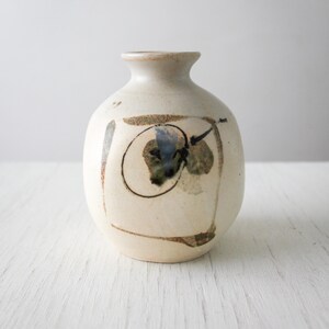 May include: A small, round, off-white ceramic vase with a narrow neck and a wide, bulbous body. The surface features a hand-painted, abstract design consisting of a thin black circular outline, brown brushstrokes, and a central blue-grey accent.