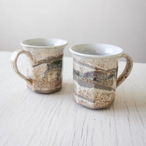 May include: Two ceramic mugs with a white glaze and brown, grey, and blue abstract patterns. The mugs have a rounded shape and a handle.