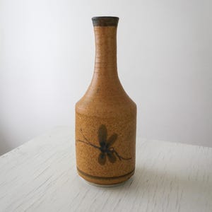 May include: A tall, slender ceramic bottle with a speckled tan and brown finish. The bottle features a narrow neck, a dark rim, and a hand-painted dark brown dragonfly motif on the lower body, accented by a thin horizontal line near the base.