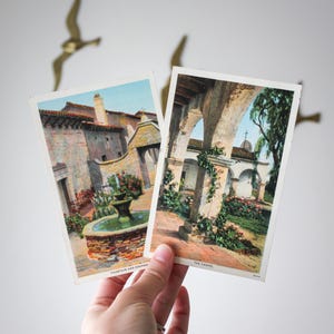 May include: Two vintage postcards featuring architectural scenes. One shows a fountain and doorway, the other depicts a chapel. The postcards are held in a hand, with text visible on each. Gold-colored bird decorations are in the background.