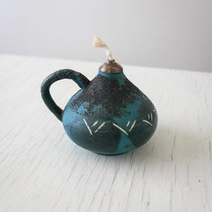 May include: A small, teal-colored oil lamp with a handle. The lamp has a dark, textured top and white markings. A silver-colored cap holds a white wick. The lamp is on a white surface.