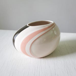 May include: A white ceramic vase with a pink and black swirl design. The vase is oval shaped and has a smooth, glossy finish.