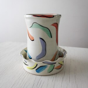 May include: A white ceramic vase and dish set. The vase is cylindrical with colourful abstract shapes in red, green, blue, and orange. The dish has a scalloped edge and similar painted designs. A decorative and functional pottery set.
