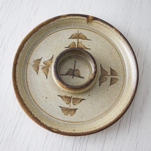 May include: A circular, beige ceramic serving platter with a central dip bowl. The dish has a brown rim and decorative brown leaf-like patterns. The dip bowl is beige with a brown rim and a similar leaf design.