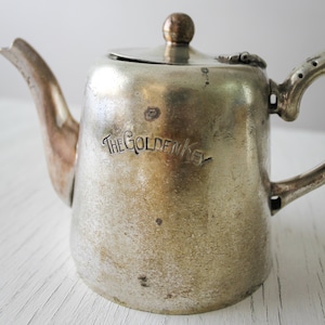 May include: A small, silver-colored teapot with a lid and handle. The teapot has the words "THE GOLDEN KEY" engraved on the front. The teapot is likely made of metal and has a vintage or antique appearance.