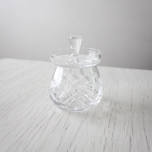 May include: A clear, cut-glass jar with a lid. The jar features a diamond pattern and a small handle on the lid. The jar is on a white wooden surface.