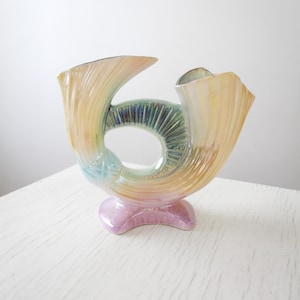 May include: A ceramic vase in the shape of a horn, with a pastel color scheme. The vase has a pink base, a central opening, and a flared top. The colors include yellow, blue, green, and pink, creating a rainbow effect. The vase is likely for decorative purposes.