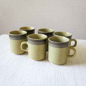 May include: Set of six green and brown striped ceramic coffee mugs. The mugs have a simple design with a band of brown around the top and bottom.