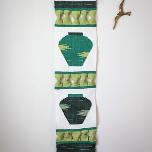 May include: A woven wall hanging with a white background, featuring two green vase designs. The top and bottom sections have green knotted accents. The wall hanging is approximately 102 cm long.