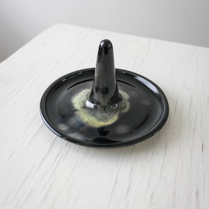 May include: A black ceramic ring holder with a cone-shaped centre and a round base. The base has a speckled design with hints of yellow and green. The ring holder is on a white surface.