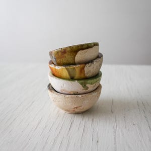 May include: Four small, handmade ceramic bowls stacked on top of each other. The bowls are in shades of beige, with colourful rims in green, yellow, and orange. They have a rustic, textured appearance, suggesting a natural, earthy aesthetic.