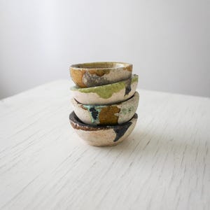 May include: A stack of four small, round, ceramic bowls with a textured, off-white base. Each bowl features irregular, hand-applied glaze patches in shades of mustard yellow, sage green, turquoise, and dark charcoal, creating a unique, mottled appearance.