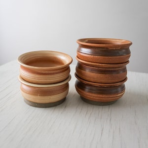 May include: A set of six small ceramic bowls with a rounded shape and a flared rim. The bowls feature a warm color palette of tan, beige, and reddish-brown tones with horizontal banding. They are arranged in two stacks, one with two bowls and one with four.