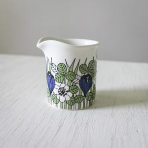 May include: White ceramic creamer with a blue and green floral pattern. The pattern features blue flowers, white flowers, and green leaves.