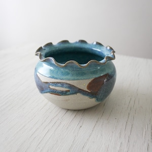 May include: A small, round ceramic bowl with a frilled rim. The bowl has a light blue, dark blue, brown, and white colour scheme. The exterior has a painted design, and the rim is a darker shade of blue.