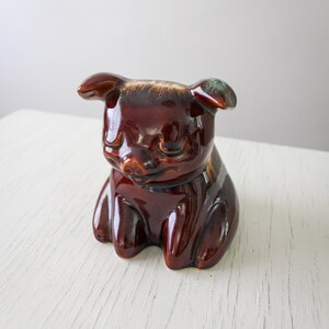 May include: A glossy, brown ceramic pig ornament with a rounded design. The pig has small ears and a smiling expression. The ornament is a decorative item, suitable for a home or office.