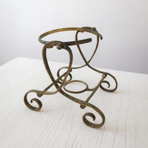 May include: A vintage brass-coloured metal stand with a circular top and decorative, scrolled legs. The stand features an open design with curved supports and a small ring near the base. The metal has an aged appearance.