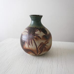 May include: A ceramic vase with a round body and a narrow neck. The vase features a brown palm tree design on a tan background. The rim of the vase is dark green.