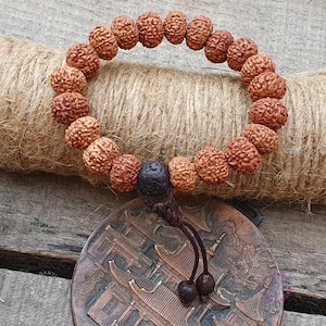 Può includere: Un braccialetto di perline mala in legno marrone con un nappa marrone scuro. Le perline sono rotonde e hanno una superficie testurizzata. Il braccialetto è appoggiato su un pezzo di stoffa di juta marrone.