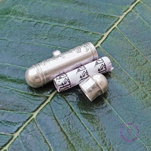 Może przedstawiać: Srebrny amulet z zwiniętym papierem w środku. Amulet jest ozdobiony misternymi wzorami, a papier pokryty jest pismem. Amulet leży na zielonym liściu.