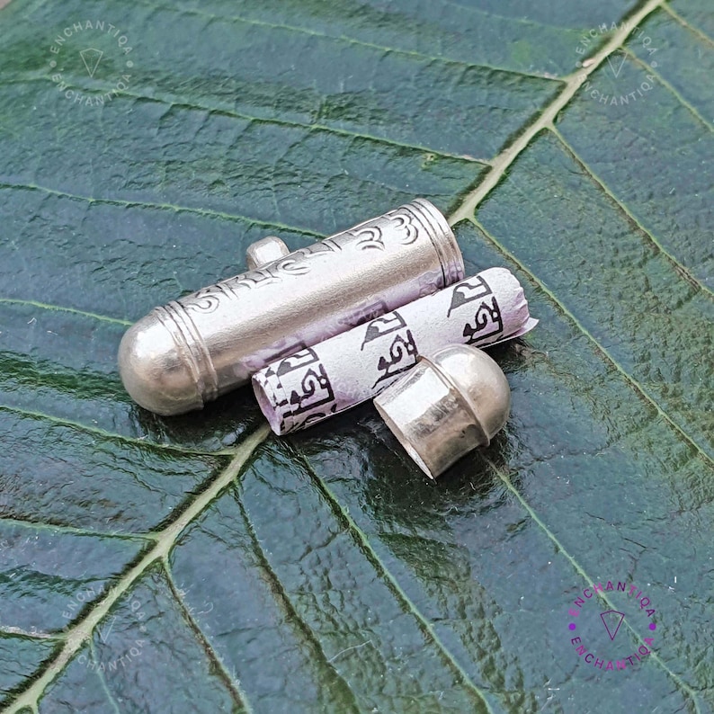Może przedstawiać: Srebrny amulet z zwiniętym papierem w środku. Amulet jest ozdobiony misternymi wzorami, a papier pokryty jest pismem. Amulet leży na zielonym liściu.