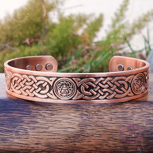 May include: A copper bracelet with a Celtic knot design and two circular medallions. The bracelet is magnetic and has a dark brown finish.