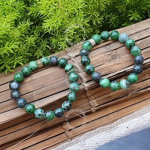 May include: Two green gemstone bracelets with silver and gold accents. The bracelets are made of round beads and are lying on a wooden surface.