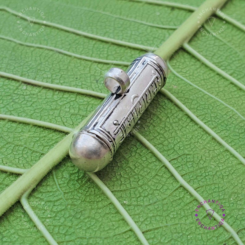 Może przedstawiać: Srebrny amulet w kształcie tuby z misternymi grawerowanymi wzorami. Zawieszka ma małą pętelkę u g&oacute;ry do przymocowania do łańcuszka.