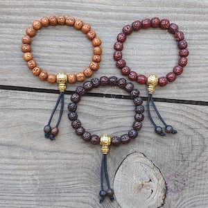 Könnte beinhalten: Drei Holzgebetperlenarmbänder mit goldenen Akzenten. Die Armbänder sind braun, rotbraun und hellbraun. Jedes Armband hat einen Quasten mit einer goldenen Perle am Ende.