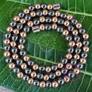 May include: A necklace composed of alternating rows of metallic gray and rose gold beads. The beads are spherical and strung together, forming a circular shape. The necklace is displayed on a green leaf.