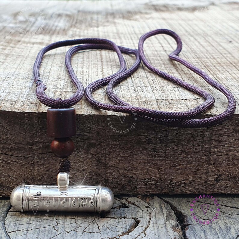 Może przedstawiać: Srebrny wisiorek amulet z ciemnobrązową drewnianą koralikiem i ciemnofioletowym sznurkiem. Amulet jest ozdobiony tekstem w obcym języku.