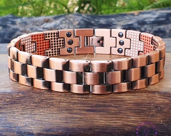 May include: A copper-colored bracelet with a geometric design. The bracelet features a series of rectangular links and decorative sections with black accents. The bracelet is displayed on a wooden surface.