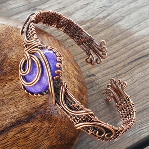 Pode incluir: Uma pulseira artesanal de fio de cobre com uma pedra preciosa roxa grande e redonda. O fio é intrincadamente tecido e moldado, com redemoinhos decorativos e extremidades abertas. A pulseira foi projetada para ser usada no pulso.