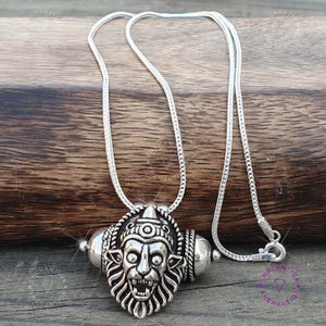 May include: A silver-colored pendant necklace featuring a detailed lion-like face with intricate carvings. The pendant is attached to a delicate chain. The necklace is displayed on a wooden surface.
