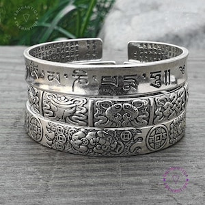 May include: Three silver-colored cuff bracelets stacked together. The top bracelet has etched characters. The middle bracelet features a repeating pattern of a dragon-like creature. The bottom bracelet has a turtle and coin design.