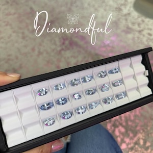 Op de afbeelding: Een zwarte displaycase gevuld met meerdere lichtblauwe edelstenen in verschillende vormen. De edelstenen zijn geplaatst in individuele witte compartimenten. Het woord "Diamondful" staat in cursief schrift over de afbeelding.