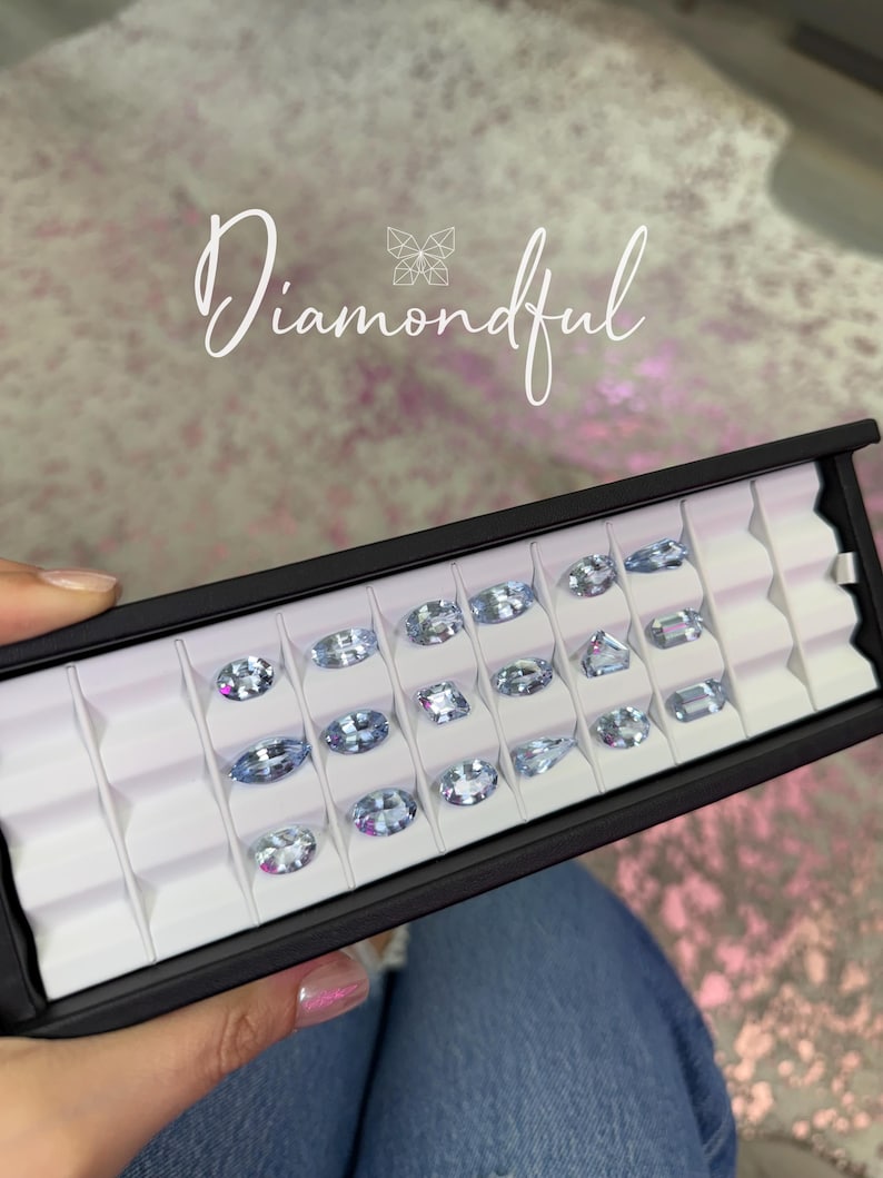 Op de afbeelding: Een zwarte displaycase gevuld met meerdere lichtblauwe edelstenen in verschillende vormen. De edelstenen zijn geplaatst in individuele witte compartimenten. Het woord "Diamondful" staat in cursief schrift over de afbeelding.