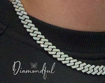 May include: A silver chain necklace with a diamond-like pattern. The chain is thick and has a chunky, boxy design. The necklace is worn by a person, but the person's face is not visible.